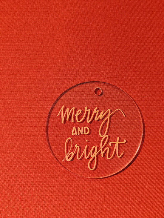 "Merry and bright" written in red ink on a clear acrylic ornament.